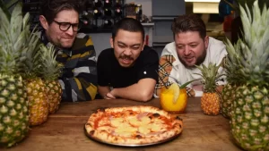 Three men look at a pineapple on pizza.