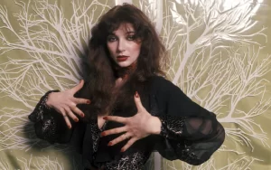 Young Kate Bush posing in an all black, sheer clothes against a ornate white and beige background